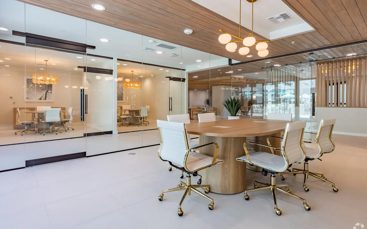 A modern conference room featuring a large round wooden table surrounded by white chairs with gold accents, glass walls, and stylish lighting fixtures.