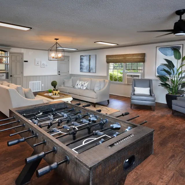 A spacious living room featuring a foosball table in the center, surrounded by a sofa, armchairs, and decorative plants, with a warm wood floor and natural light coming through the windows.