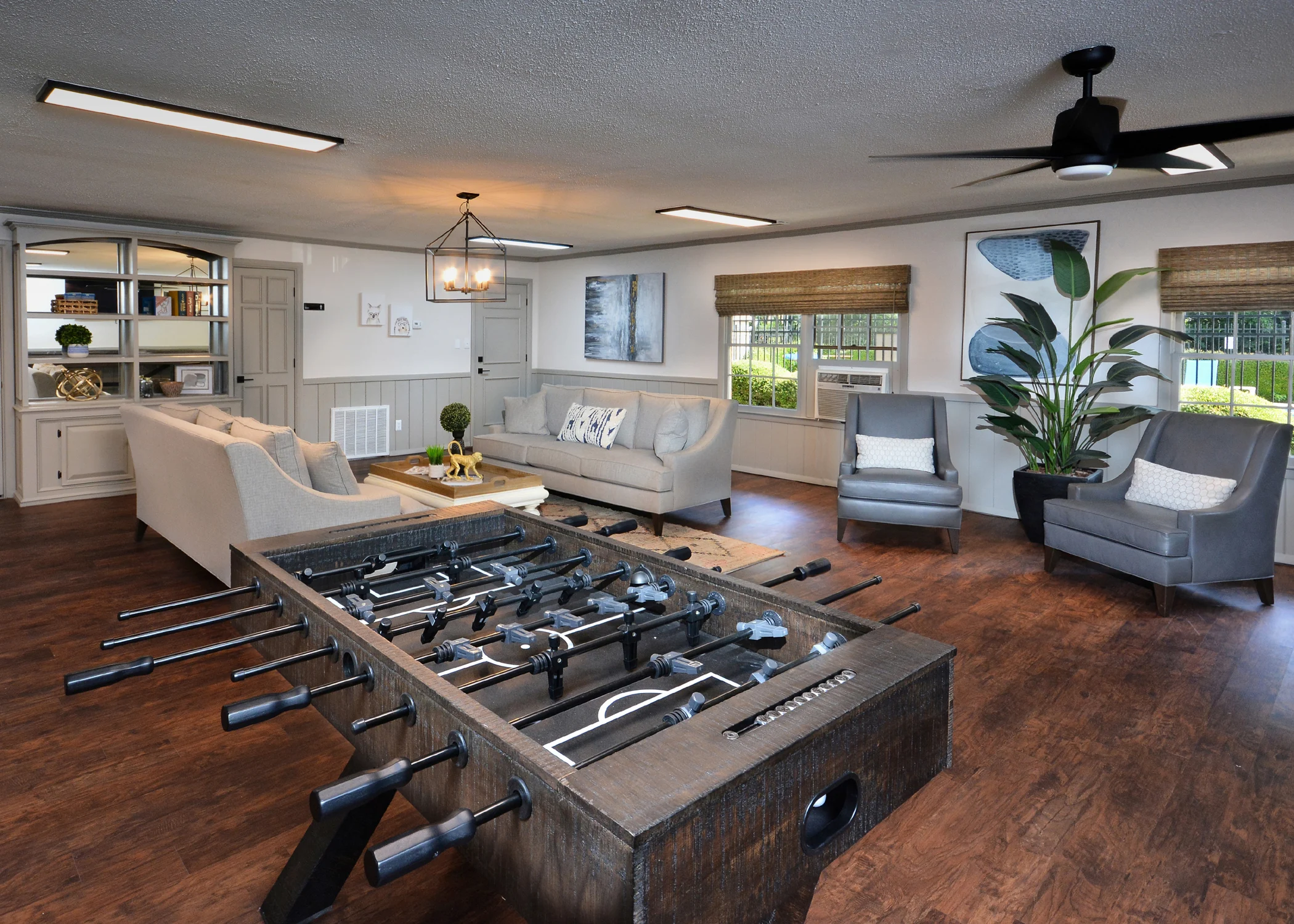 Spacious Living Room with Foosball Table A spacious living room featuring a foosball table in the center, surrounded by a sofa, armchairs, and decorative plants, with a warm wood floor and natural light coming through the windows.