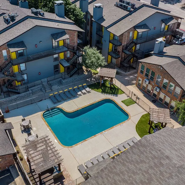 Aerial view of an apartment complex featuring a swimming pool, surrounding buildings, and parking area.