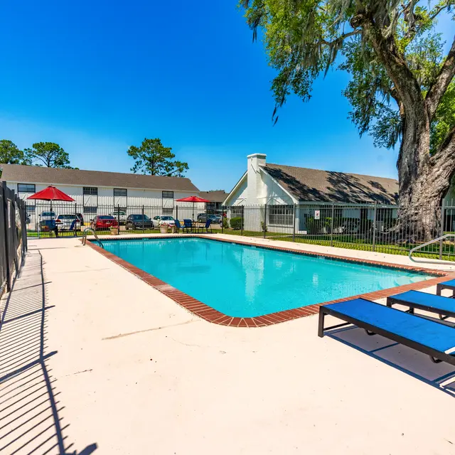 A bright outdoor swimming pool surrounded by a fence, with blue lounge chairs on one side and a shaded area with umbrellas in the background.
