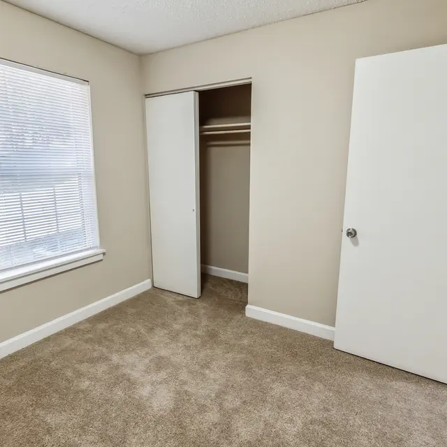 An empty room with beige walls and carpet, featuring a window with blinds and an open closet.