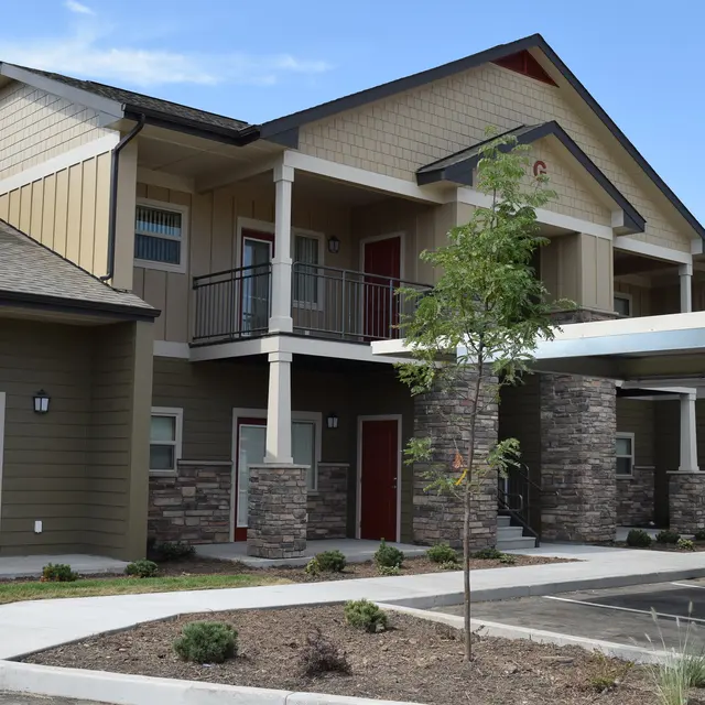 Stylish Modern Apartment Building A modern apartment building featuring a mix of stone and wooden siding, with a crisscross design, green and beige colors, and a balcony. A pathway leads to the entrance, and there are well-landscaped areas with small bushes.