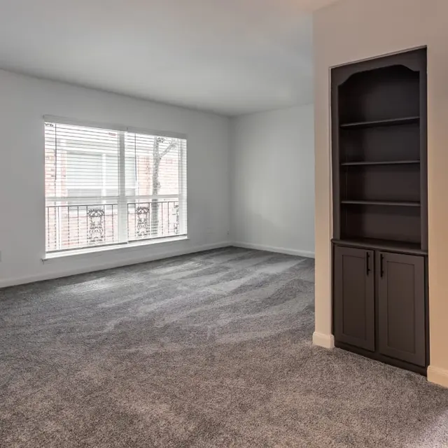 Empty living room with carpet flooring, a window with blinds, and a built-in shelf.