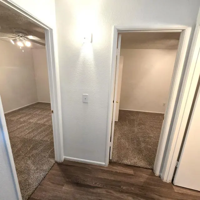 Hallway with Doorways to Rooms A view of a hallway with three doorways leading to rooms. The hallway has beige carpeting and wood flooring visible. The left room has a ceiling fan and is well lit, while the right room leads to a light, empty space. The walls are painted white.