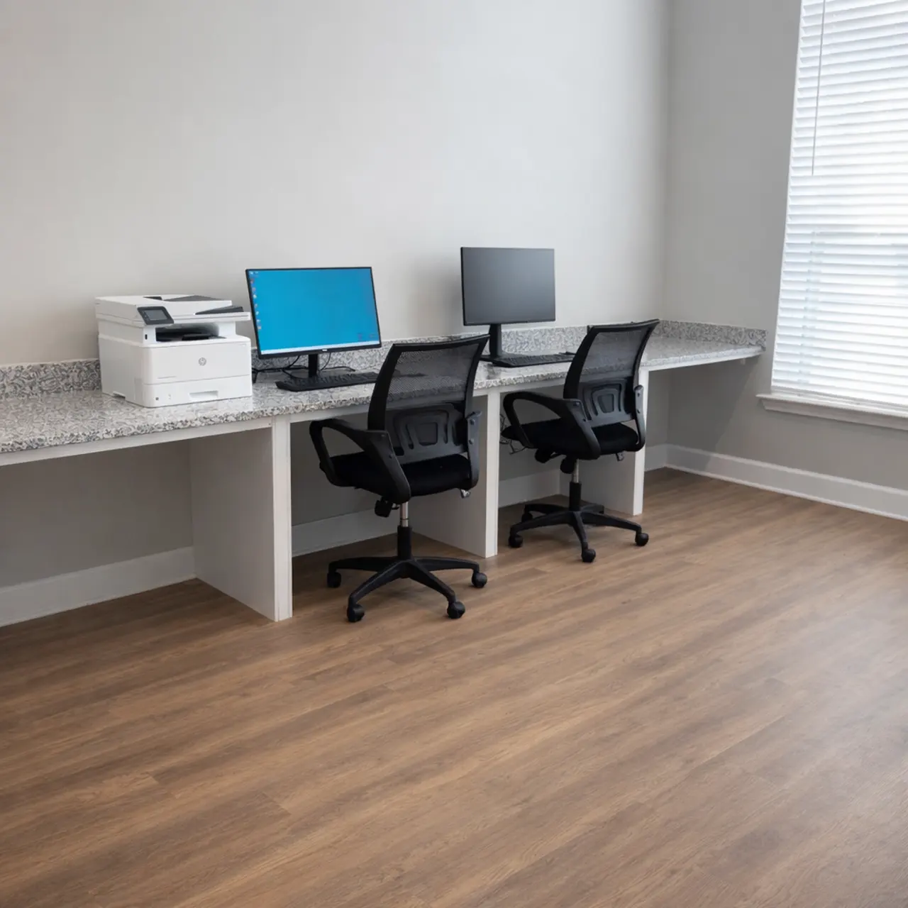 A modern office workspace featuring two desks with ergonomic chairs, each equipped with computer monitors. A multifunction printer is placed on the counter, and there are large windows letting in natural light.