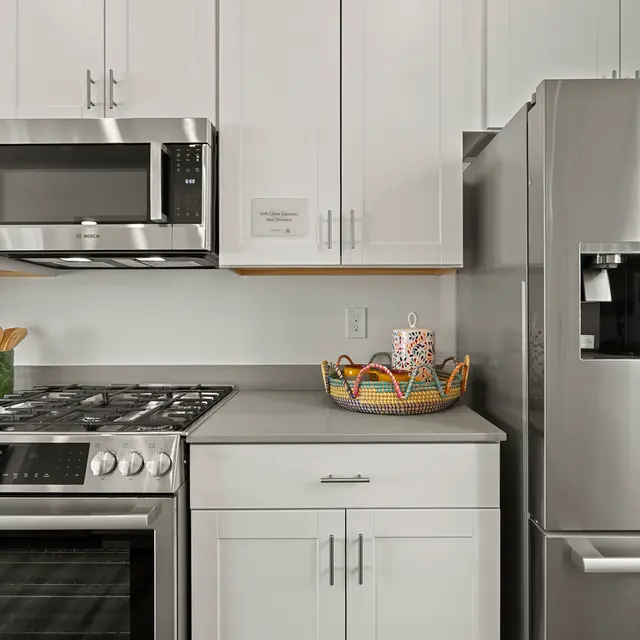 A modern kitchen featuring a stainless steel microwave, oven, and refrigerator, all with white cabinetry and a countertop displaying a decorative basket with kitchen items.