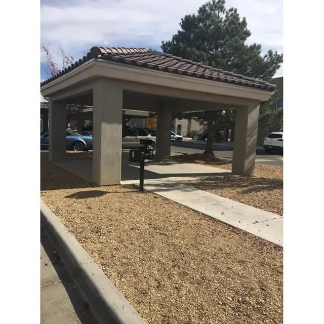 A covered outdoor structure with a tiled roof, set in a gravel area with a paved path. Surrounded by trees and parked cars in the background.