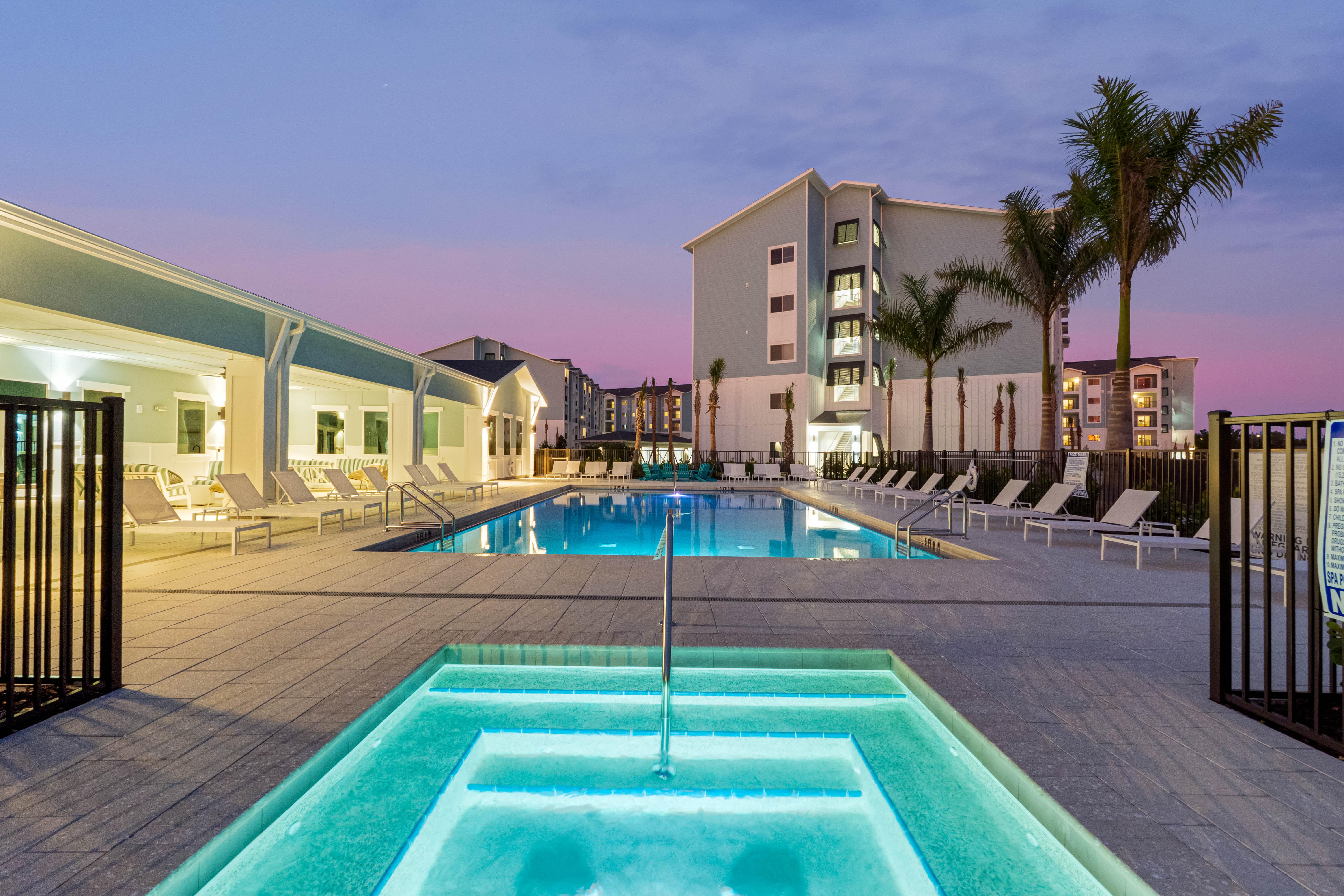 A modern swimming pool area with two pools, lounge chairs around the poolside, and palm trees, set against a colorful twilight sky.