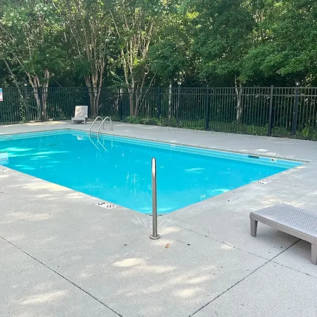 An outdoor swimming pool with clear blue water, surrounded by a concrete deck and a fence. A lounge chair is placed beside the pool.