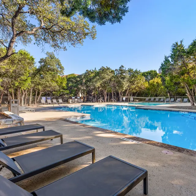 A serene swimming pool area surrounded by trees, featuring lounge chairs and clear blue water under a clear sky.
