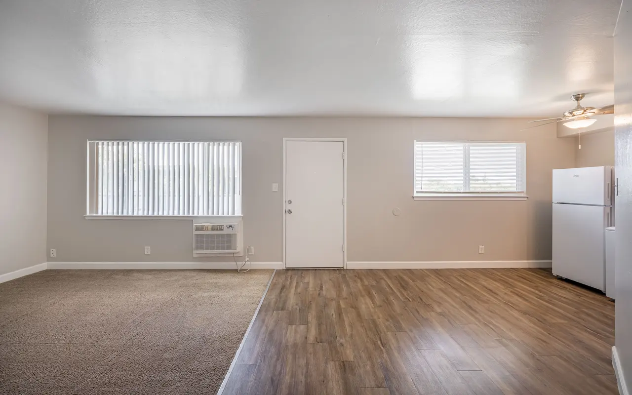 A spacious living room featuring a combination of carpet and wooden flooring, with large windows allowing natural light. A door leads to the outside, and a refrigerator is located to the right.