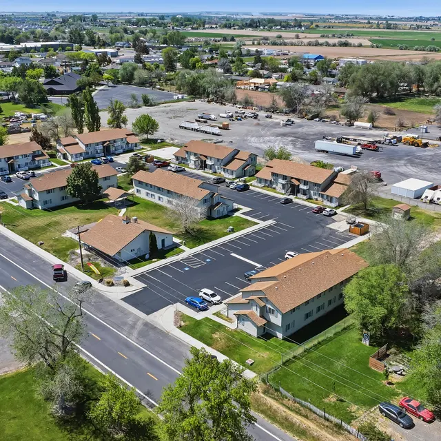 Aerial View of Suburban Area and Fields Aerial view of a suburban area featuring several residential buildings with orange roofs, a newly paved parking lot, and a variety of trees. In the background, agricultural fields stretch out, and a larger parking lot for trucks is visible.