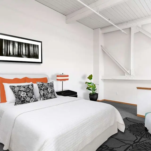 A modern bedroom featuring a white bed with patterned pillows and orange accents, black nightstands with orange lamps, a plant, and a cozy chair on a patterned rug.