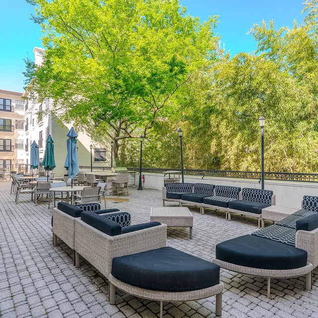 A spacious outdoor lounge area featuring black wicker sofas and chairs, a few tables under teal umbrellas, and a lush backdrop of trees.