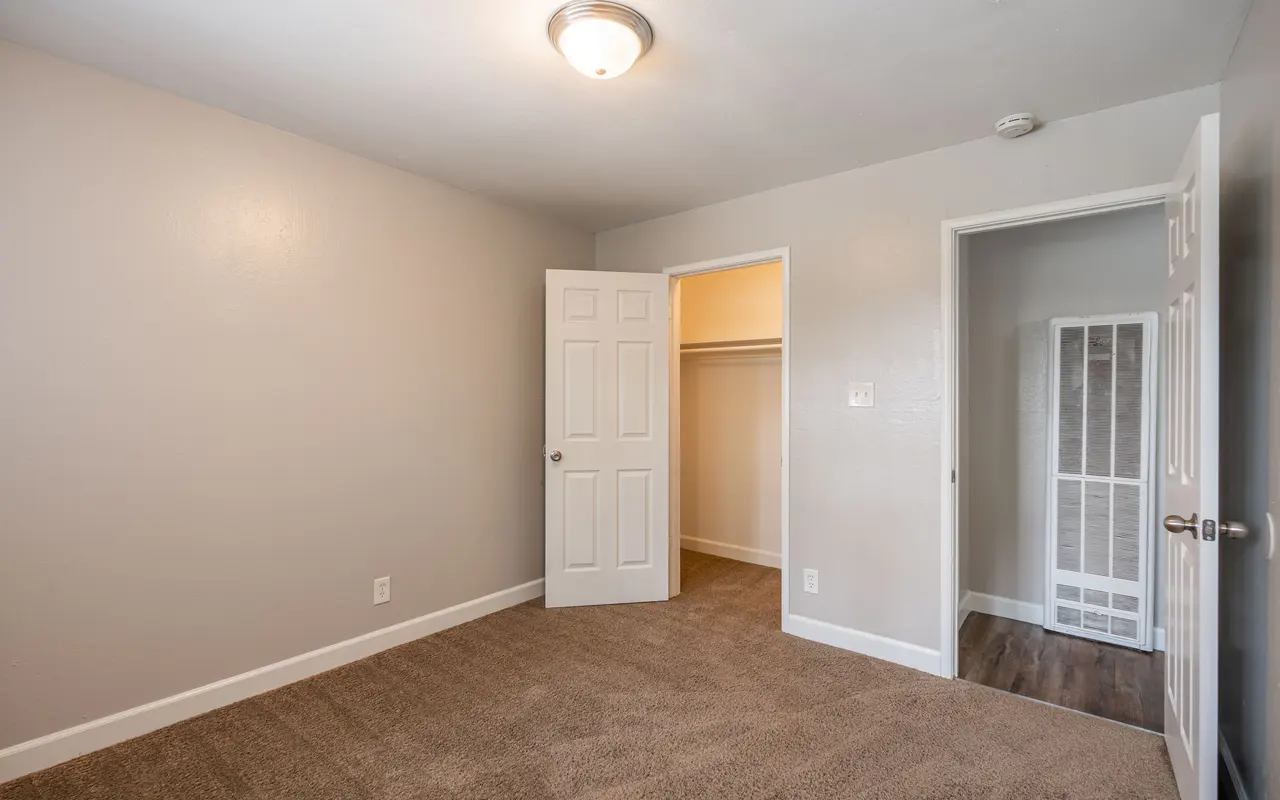 A spacious, empty room featuring a light-colored wall and a tan carpet. There are two doors: one open to a closet and another leading to the hallway. The ceiling is illuminated by a central light fixture.