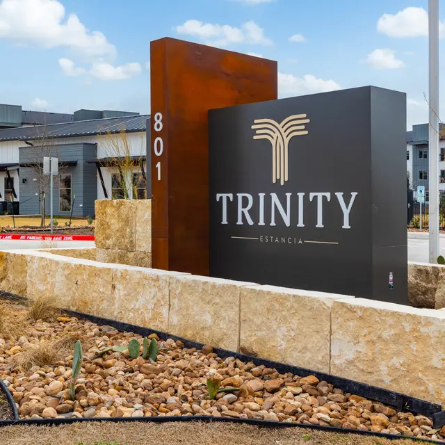 A modern sign for Trinity Estancia, set in a landscaped area with rocks and plants, featuring the text 'TRINITY' prominently displayed.