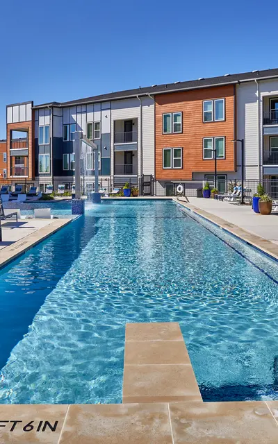 View of a modern apartment complex swimming pool area with lounge chairs and clear blue water under a sunny sky.