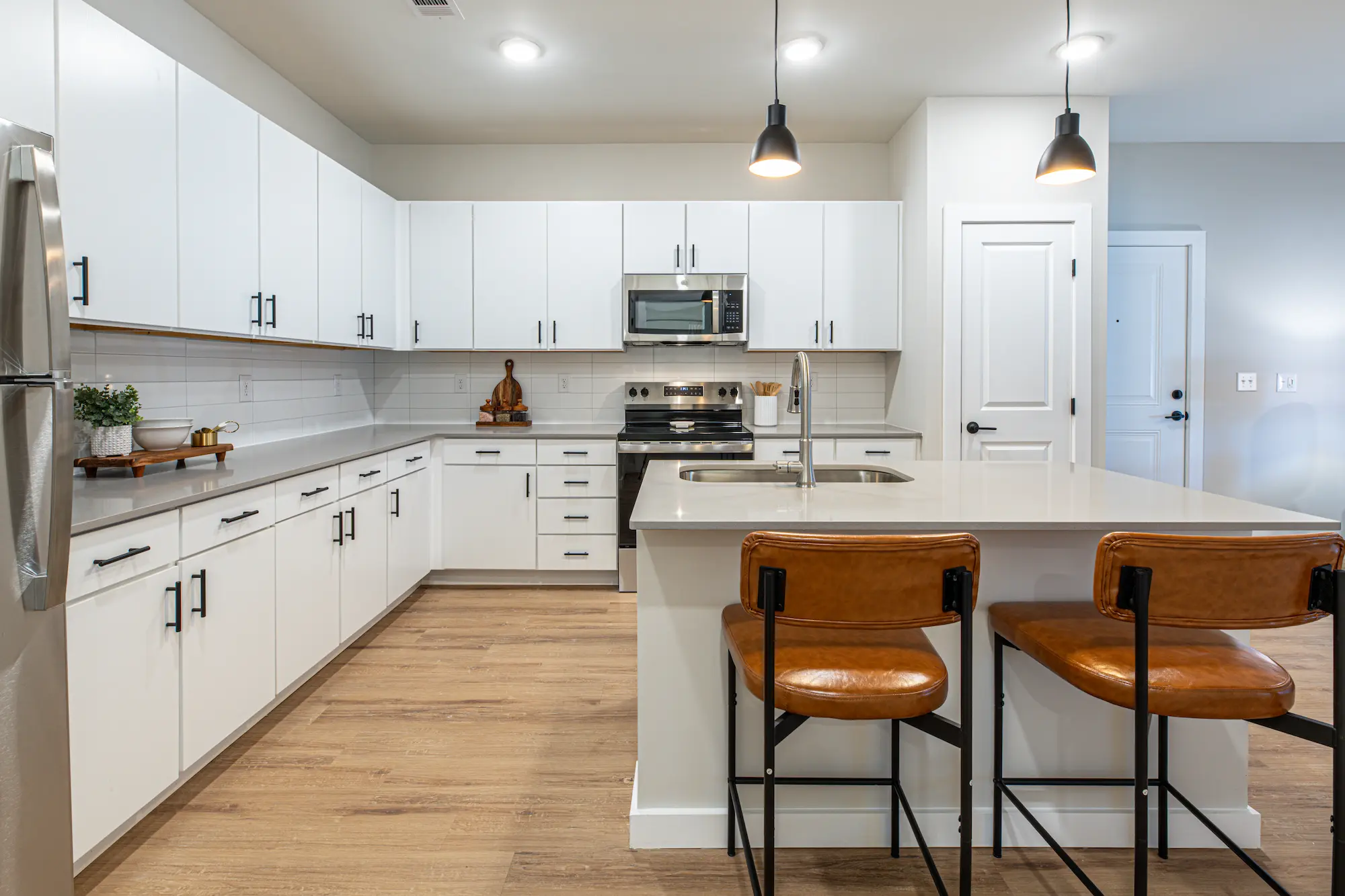 A modern kitchen featuring white cabinetry, stainless steel appliances, and a central island with seating. The kitchen has wooden flooring and pendant lighting above the island.