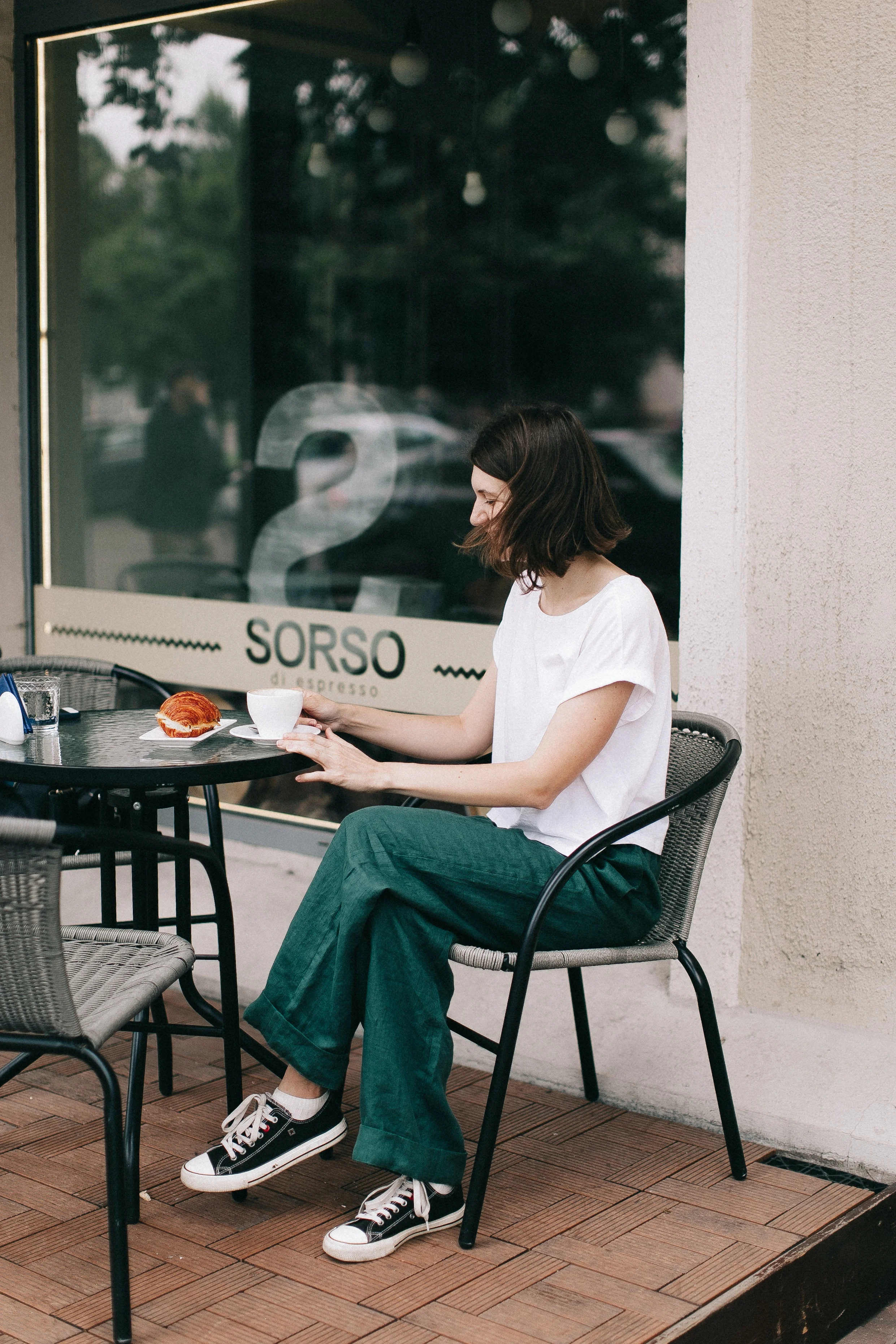 A woman sitting at a cafe table, wearing a white shirt and green pants, with a plate of food in front of her. She appears to be looking at her phone. The cafe has a large window with the name 'SORSO' visible.