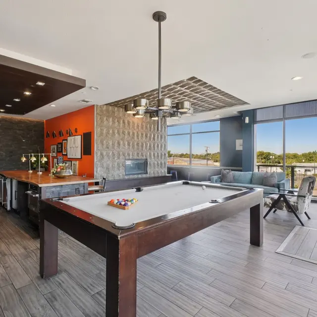 A modern game room featuring a pool table, wooden flooring, and large windows with a view. There's a bar area with seating in the background.