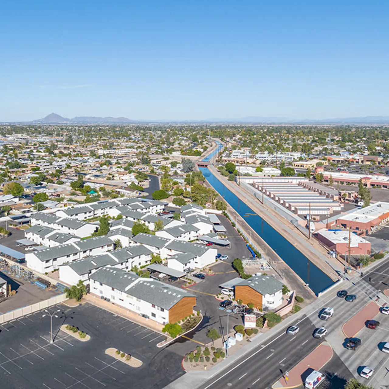 An aerial view of a suburban city with a water canal running through it. The scene includes residential buildings, commercial structures, and roads with cars. The landscape is flat with scattered greenery and mountains in the distance.