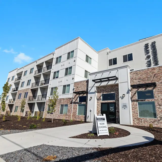 A modern apartment building with a combination of brick and light-colored siding, featuring multiple balconies. A sign in front indicates 'Now Leasing.'
