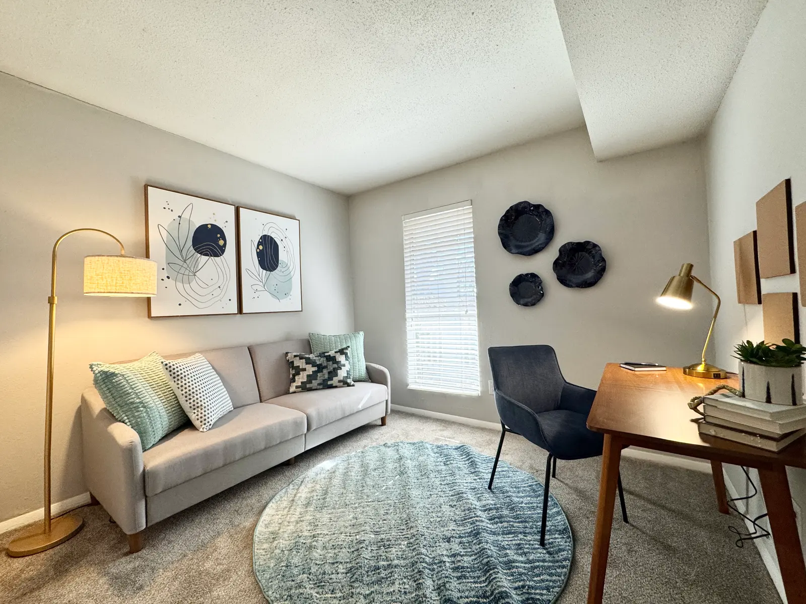 A cozy living room setup featuring a light gray sofa adorned with decorative cushions, a small round rug, a wooden desk with a desk lamp, and framed art on the wall.