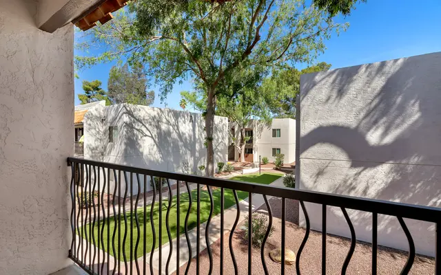 A view from a balcony overlooking a landscaped area with green grass and trees, featuring white buildings in the background.