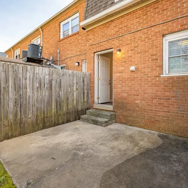 A small concrete backyard area with steps leading to a door, enclosed by a wooden fence. There is a potted plant in the corner and a grassy area in the background.