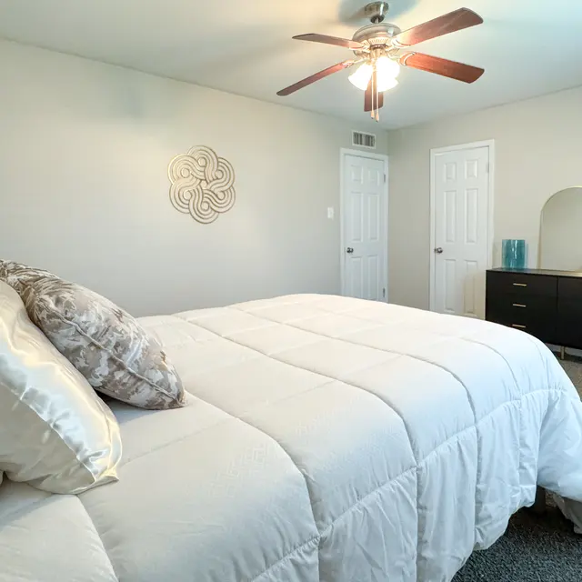 A cozy bedroom with a white bedspread, decorative pillows, a ceiling fan, and a modern dresser with a mirror.