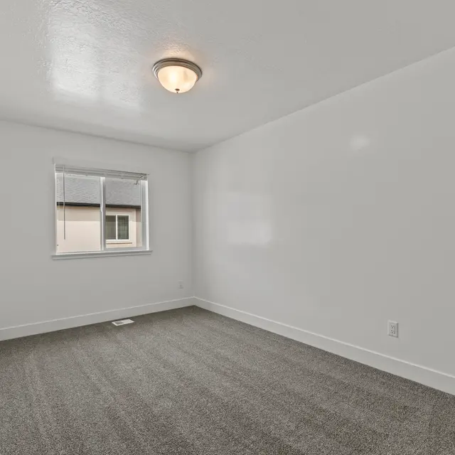 An empty room with light gray carpet and white walls. There is a window on one side and a ceiling light fixture.