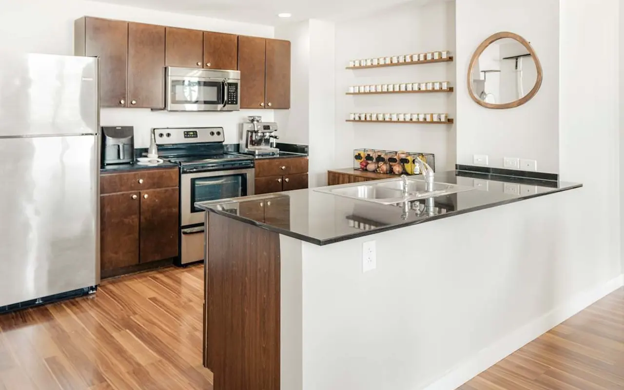 A modern kitchen featuring dark wood cabinets, stainless steel appliances, a granite countertop island with a sink, and open shelving displaying jars.