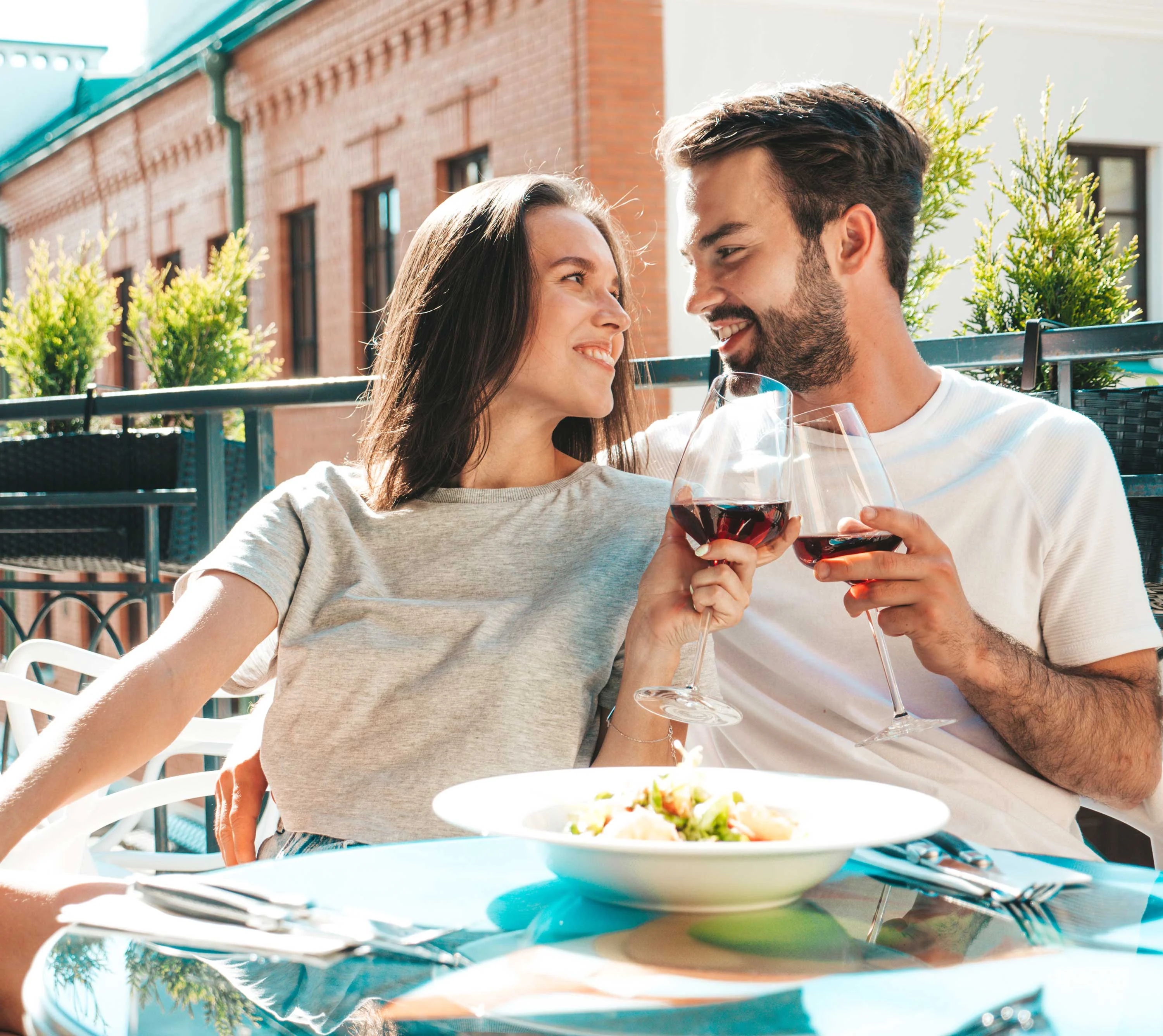 A happy couple sitting at a table outdoors, toast with wine glasses, surrounded by greenery.