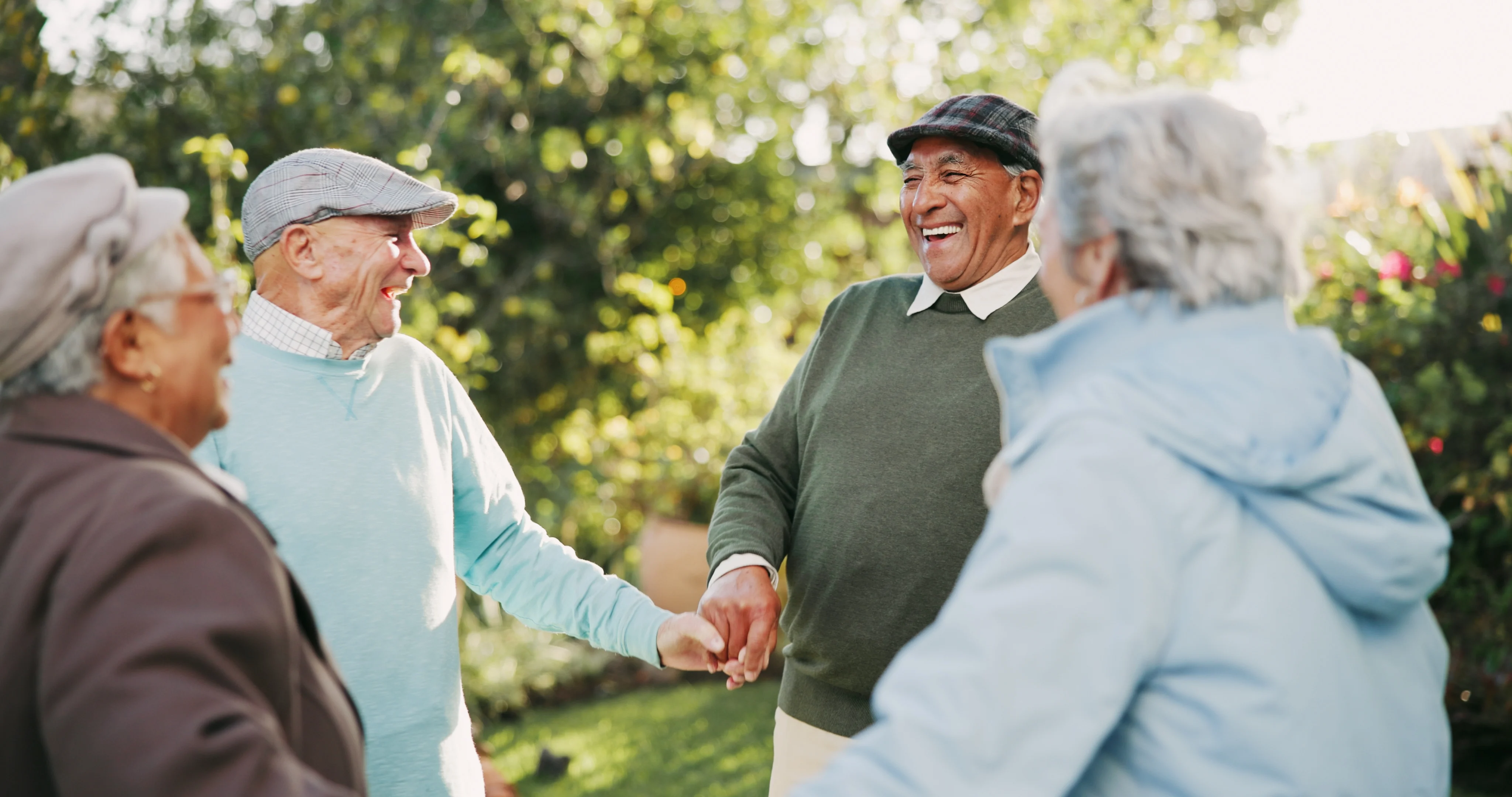 A joyful gathering of four seniors holding hands and smiling in a sunny garden. They are wearing light sweaters and caps while surrounded by greenery and flowers.