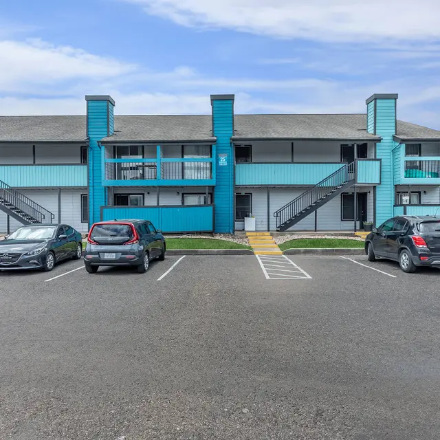 Exterior view of an apartment complex with turquoise accents, featuring multiple units, parking area, and clear blue sky.