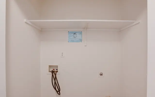 An empty laundry room space with a white shelf and utility connections on the wall.