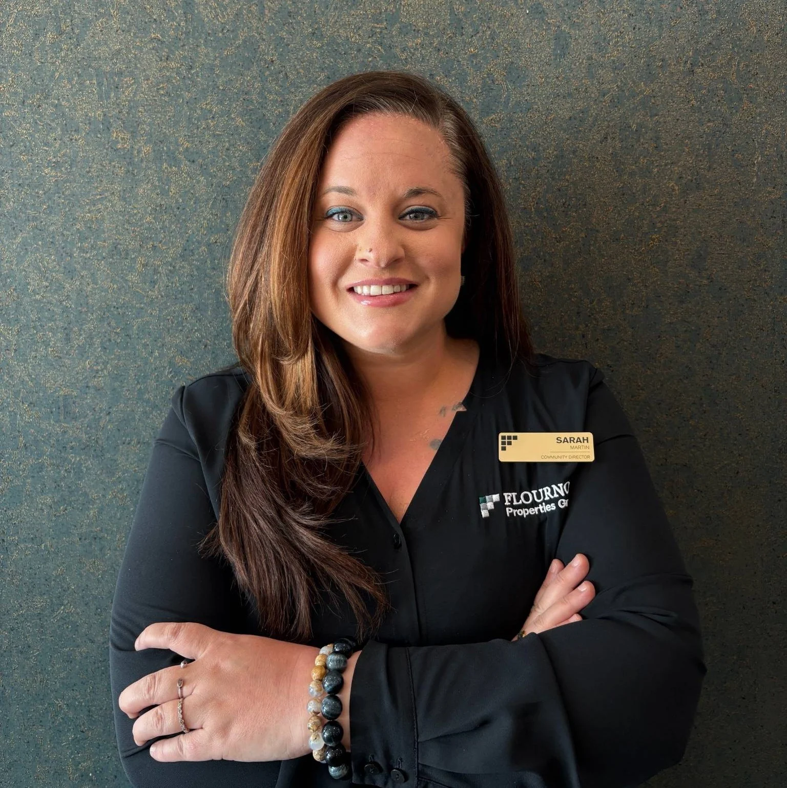 Professional Portrait of Sarah A woman with long brown hair in a black blouse, smiling with arms crossed, standing against a textured blue background. She has a name tag that reads 'SARAH' and is affiliated with 'FLOURISH Properties Group'.