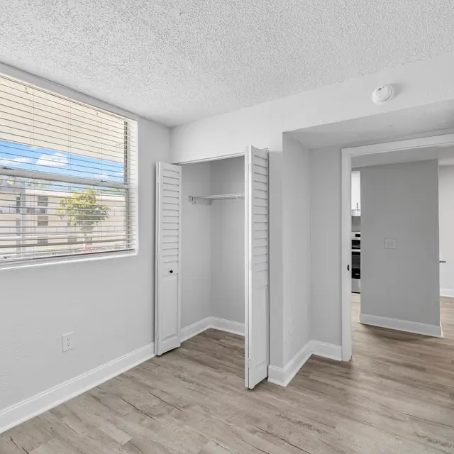 A vacant room with light wood flooring, a window with blinds, and two white doors leading to a closet and another space. The walls are painted white.
