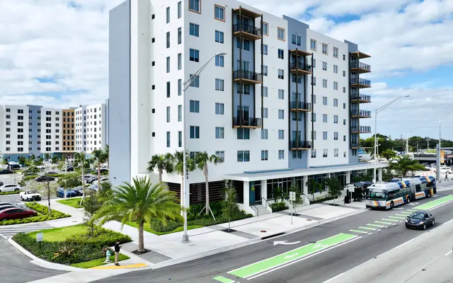 A modern multi-story apartment building with balconies, located near a busy street. The building features palm trees in the foreground and a dedicated bike lane visible on the road.