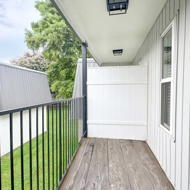 A balcony view with a wooden floor, black railings, and a white wall. In the background, there are neighboring buildings with gray and blue exteriors, and greenery is visible below.