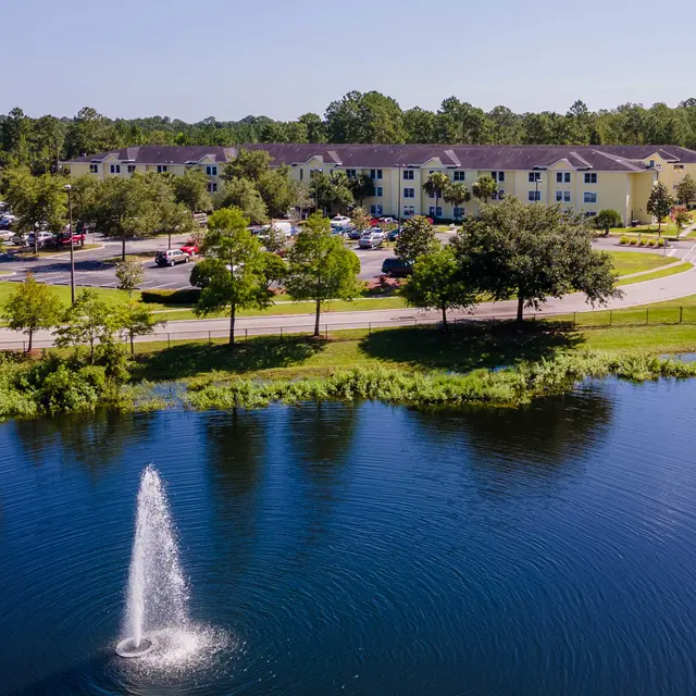 Aerial view of a hotel by a lake featuring a fountain, surrounded by greenery and parked cars.