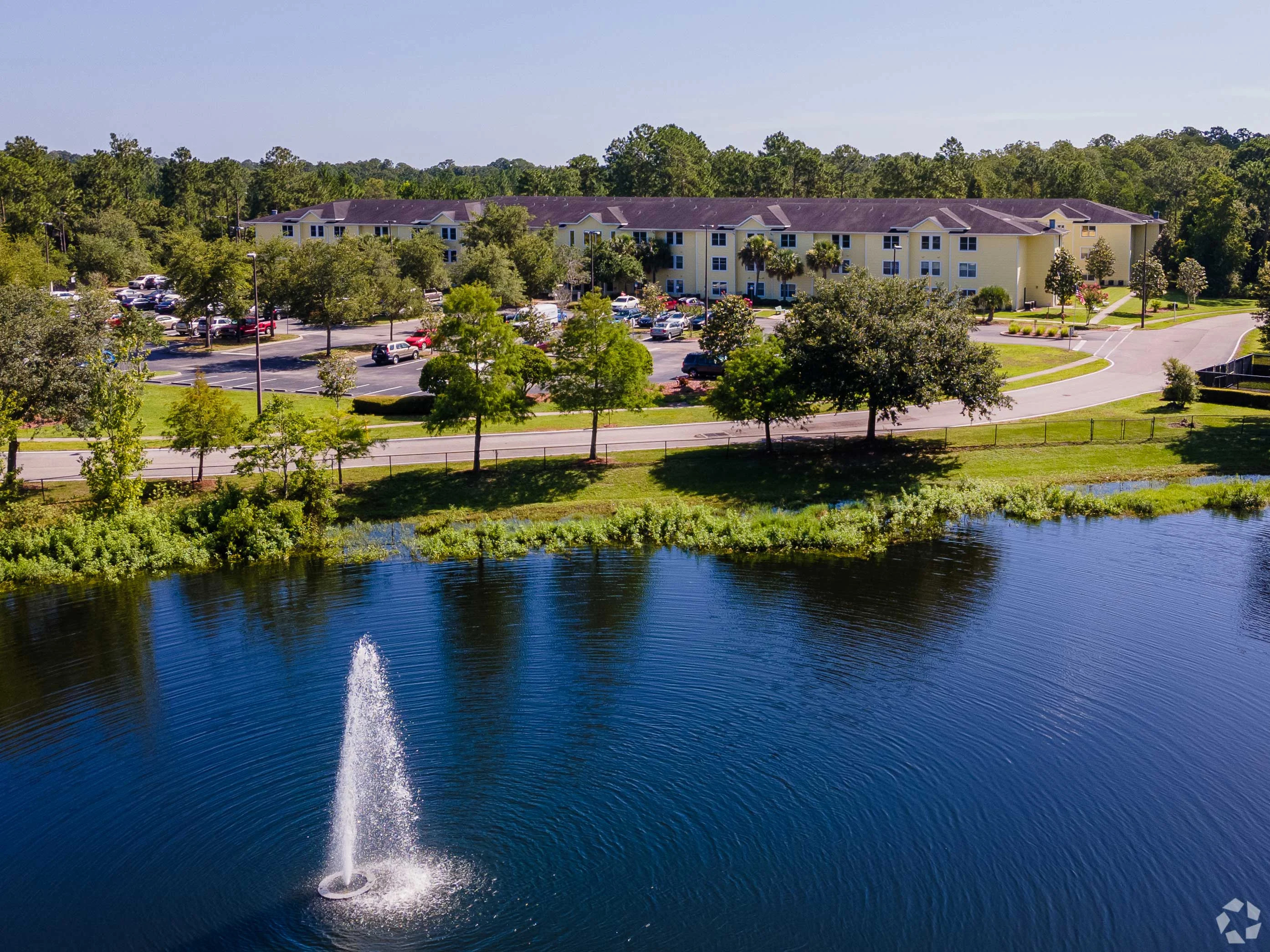 Aerial view of a hotel by a lake featuring a fountain, surrounded by greenery and parked cars.