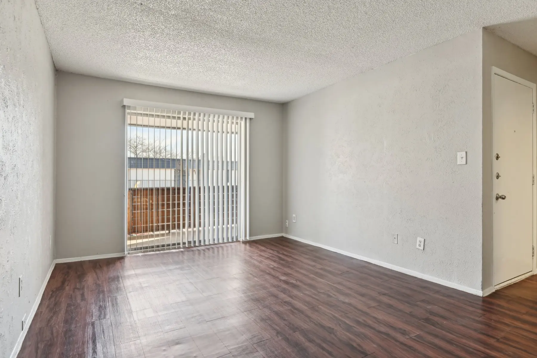 A vacant living room with laminated wooden flooring and a window covered with vertical blinds, allowing natural light to enter.