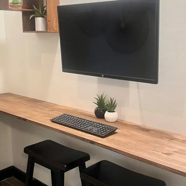 A cozy workspace featuring a mounted monitor above a wooden desk with a keyboard and small potted plants, flanked by two black stools.