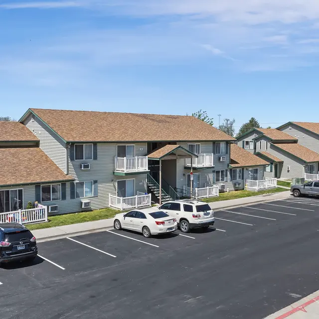 Residential Apartment Complex A row of low-rise apartments with a parking lot in front. The buildings have a combination of green siding and brown roofs, with balconies visible on the lower levels. Several cars are parked in the lot, and the surrounding area is clear with a grassy patch nearby.
