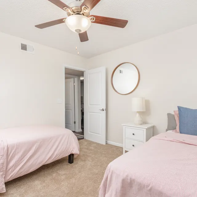 A well-lit bedroom featuring two beds with light pink bedding and blue pillows, a small nightstand with a lamp, and a round mirror on the wall.