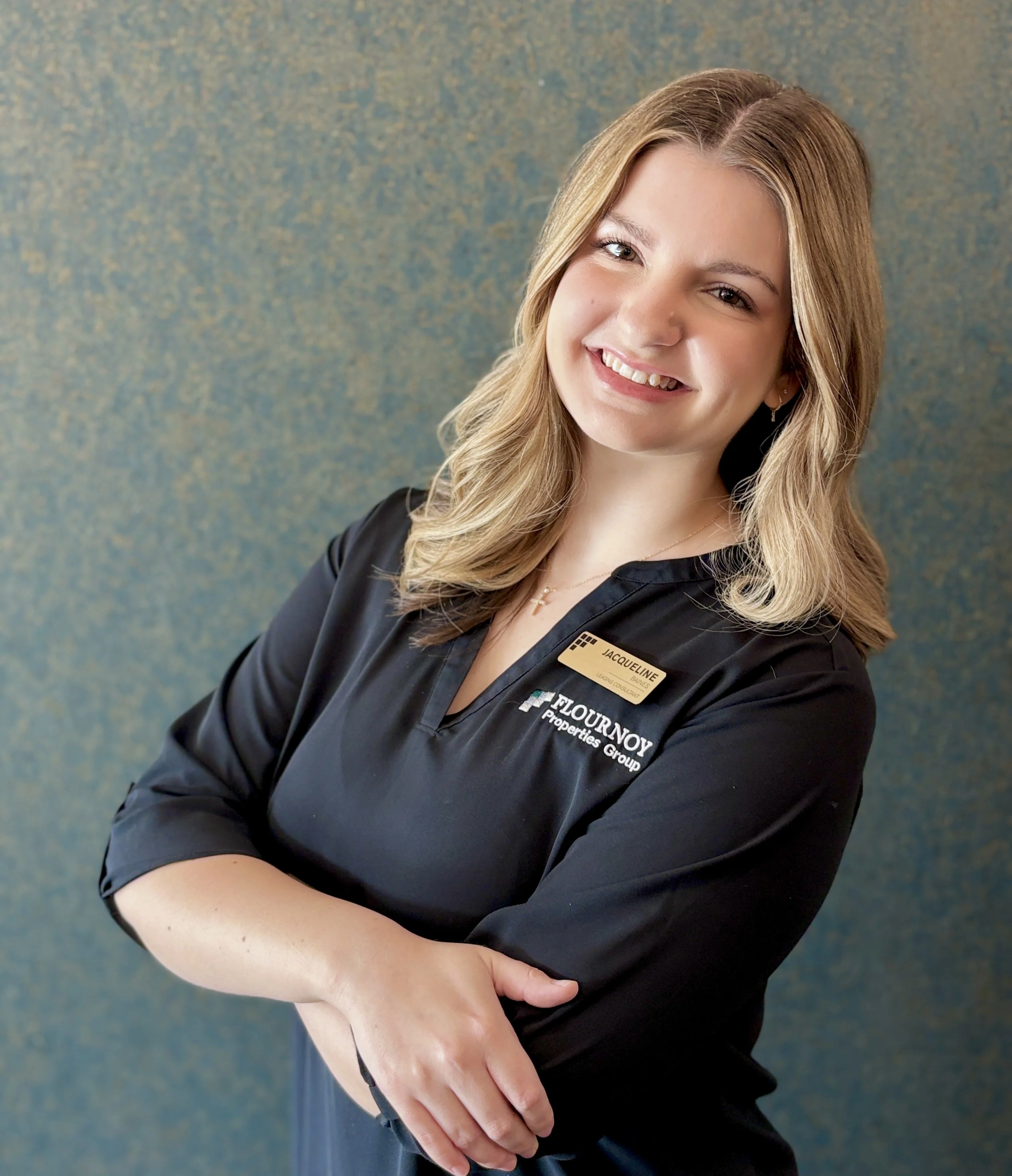 Professional Portrait of a Businesswoman A smiling woman in a black blouse with a name tag, posing confidently with her arms crossed against a textured blue background.
