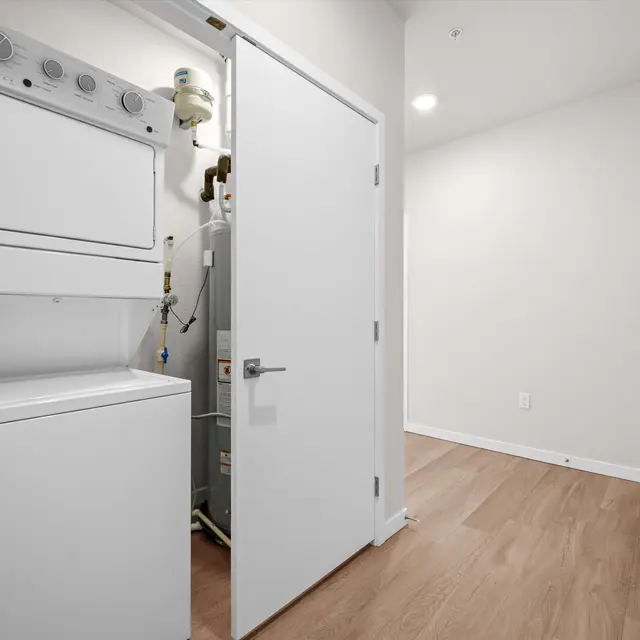 A compact laundry room featuring a stacked washer and dryer on the left, with a water heater visible behind a door on the right side. The room has light-colored walls and a wooden floor.
