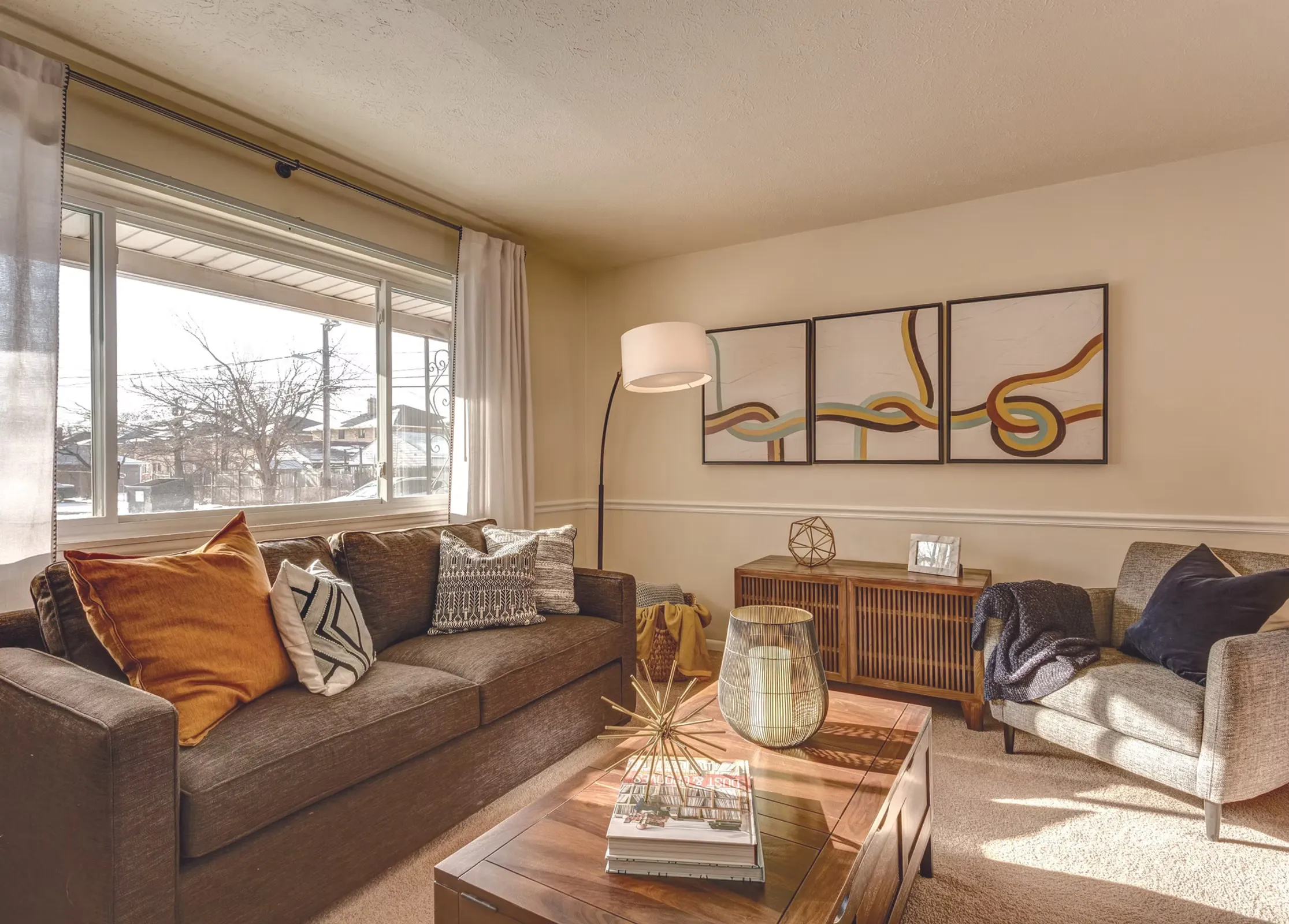A cozy living room featuring a gray sofa with colorful pillows, a wooden coffee table with decor, and a large window allowing natural light.